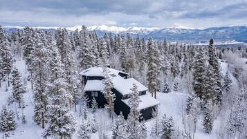 A cabin surrounded by a dense forest of snow-covered trees, with a mountain range in the background.