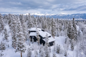 A cabin surrounded by a dense forest of snow-covered trees, with a mountain range in the background.
