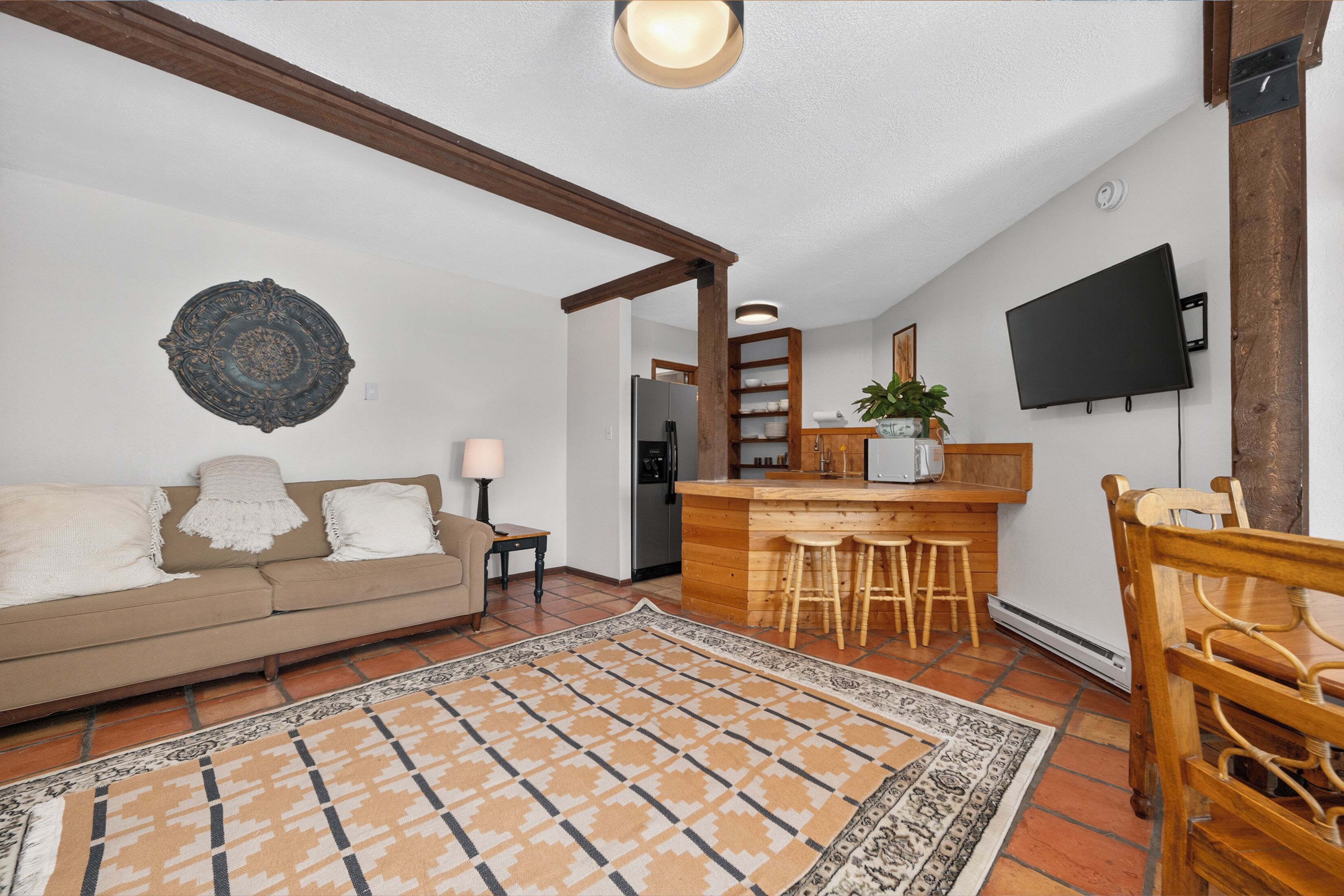 A living room with a sofa, a coffee table, a patterned rug, a wooden dining table with chairs, a kitchen area with a bar, and a flat-screen TV mounted on the wall.
