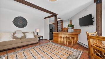 A living room with a sofa, a coffee table, a patterned rug, a wooden dining table with chairs, a kitchen area with a bar, and a flat-screen TV mounted on the wall.