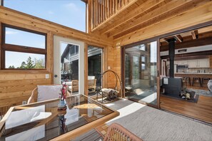 A wooden cabin interior with a glass dining table, wicker chairs, a fireplace, and a kitchen area.