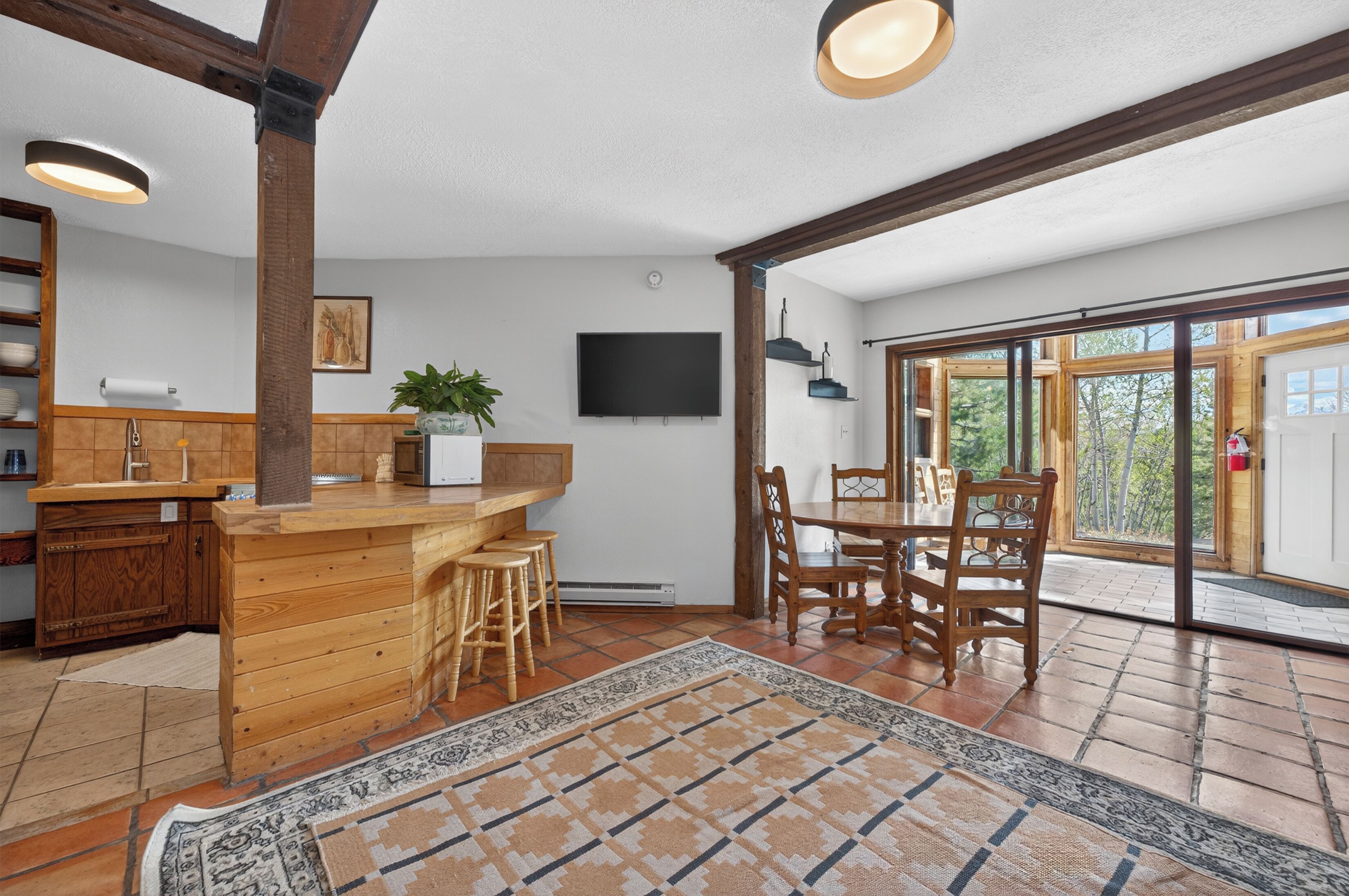 A kitchen with wooden cabinetry, a bar area, a dining table with chairs, a TV mounted on the wall, and a large sliding glass door leading to an outdoor area.