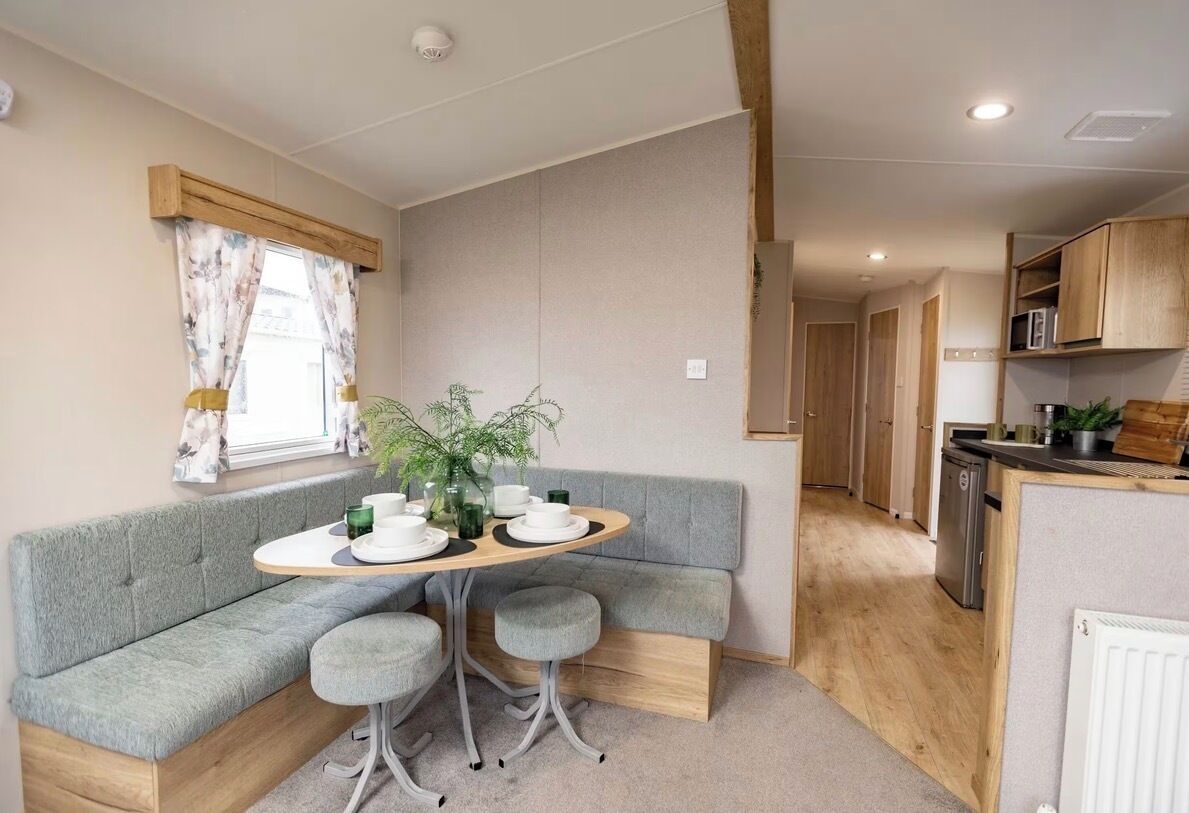 Een kleine keuken met houten kastjes, een zithoek met een grijze bank en tafel, en een raam met gordijnen.