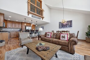 A spacious living area with a high ceiling, wooden flooring, a brown leather sofa, a coffee table with pinecones, and a dining area with wooden cabinets and a chandelier. - Summit Trail J3 views from private deck, vaulted ceilings, spacious floor plan (Fraser)