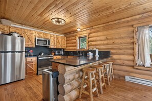 Cocina tipo cabaña de troncos con una isla central, taburetes de madera, electrodomésticos de acero inoxidable y una ventana con cortinas.