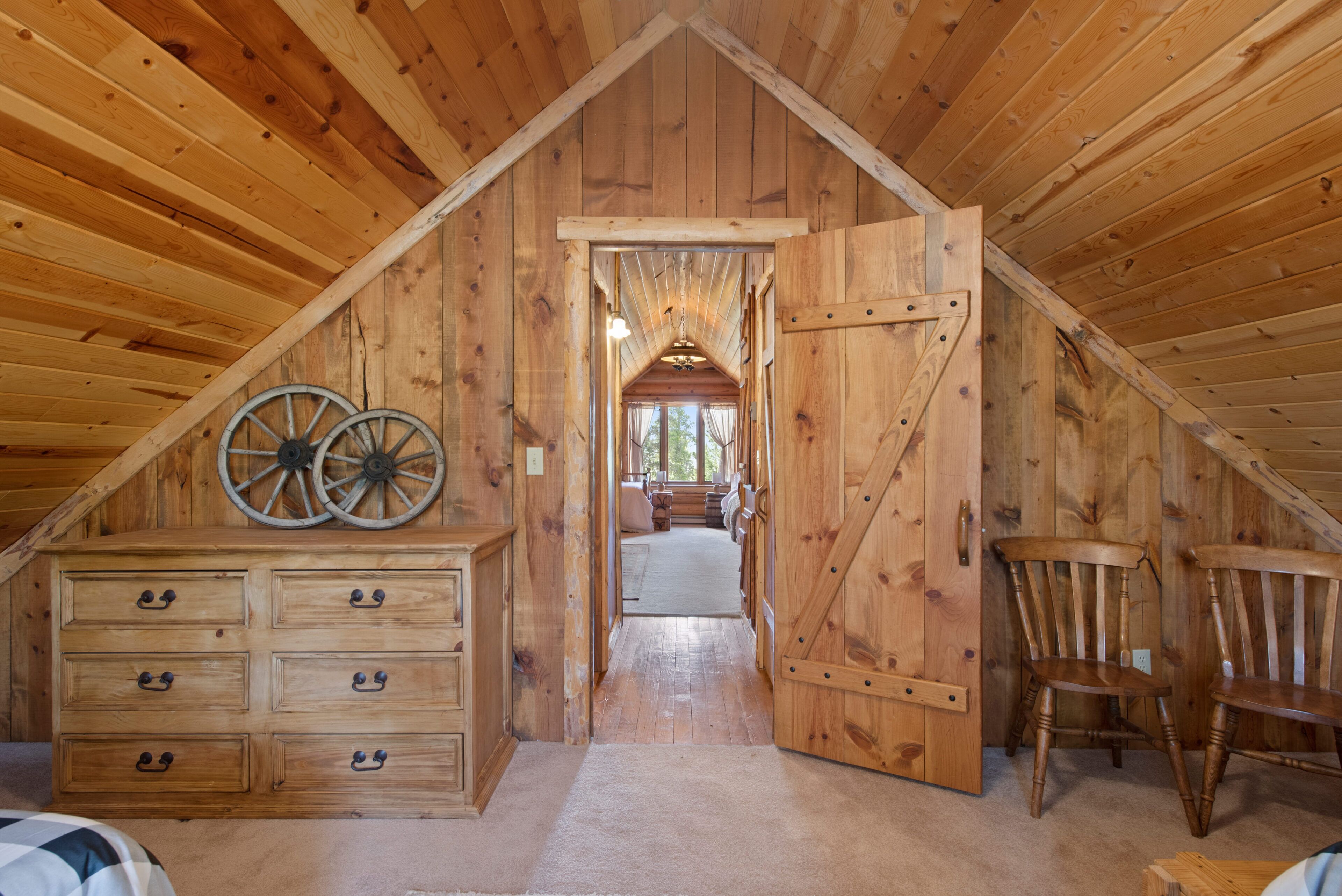 Une pièce mansardée en bois avec un plafond haut, une commode dotée de nombreux tiroirs, deux chaises en bois et une grande porte en bois.
