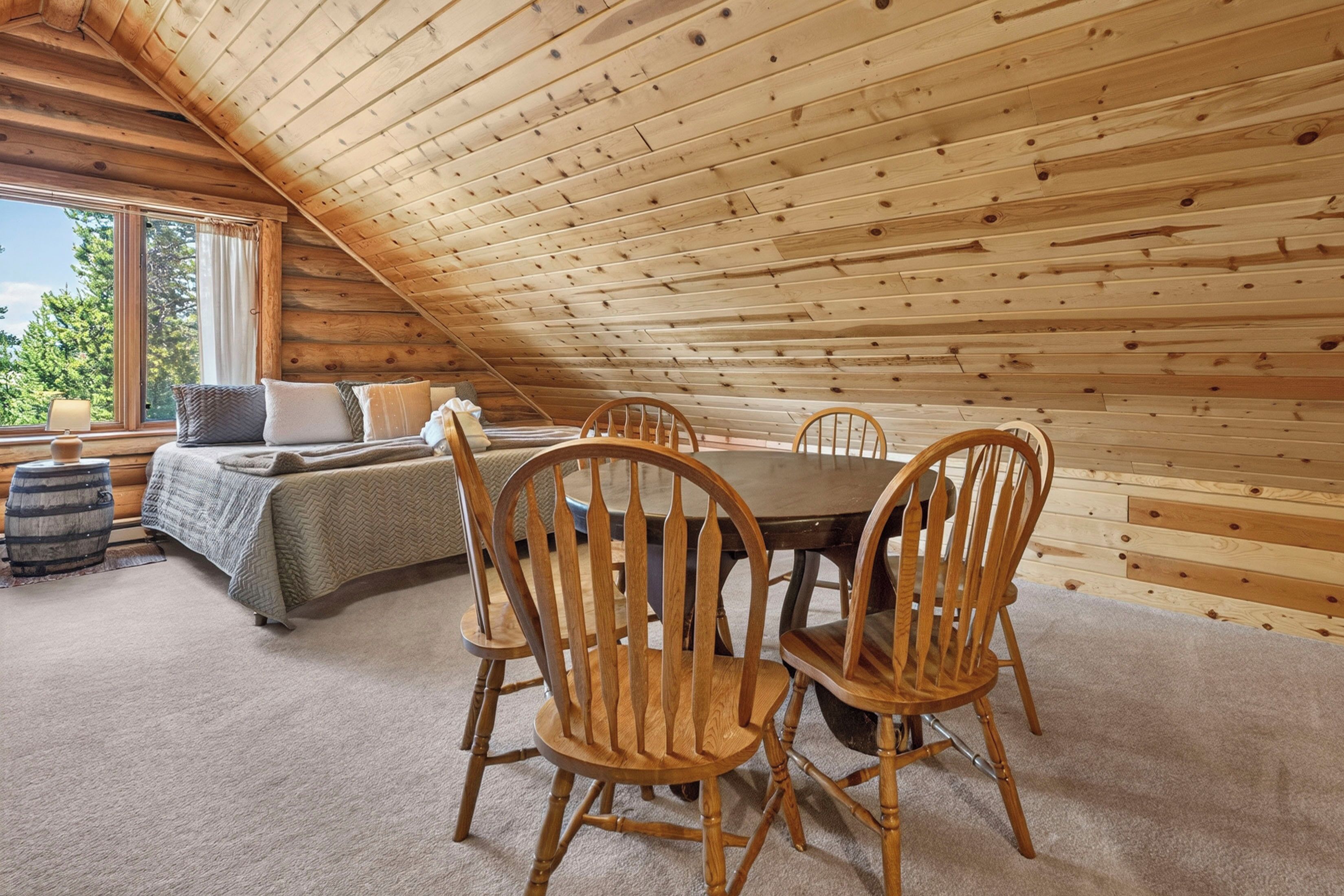 A cozy attic bedroom with a wooden sloped ceiling, a bed with pillows, a small table, and chairs.