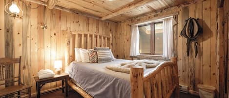 Dormitorio de cabaña de madera con cama, mesita de noche, lámpara y ventana.