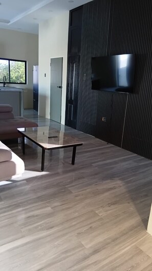 Ein modernes Wohnzimmer mit einem Flachbildfernseher an einer dunklen Wand, einem Glastisch und einem Holzboden.