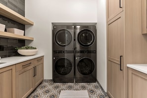 A compact laundry room with a double-door washer and dryer, wooden cabinets, a white countertop, and patterned floor tiles. - Paradise Palace: Lagoon Front! Pool + Waterslide (St. George)