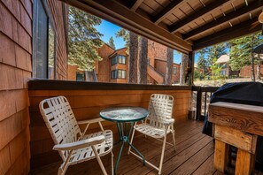 Un patio exterior con dos sillas blancas de mimbre y una pequeña mesa redonda, suelo de madera y vistas a un edificio de ladrillo con árboles.