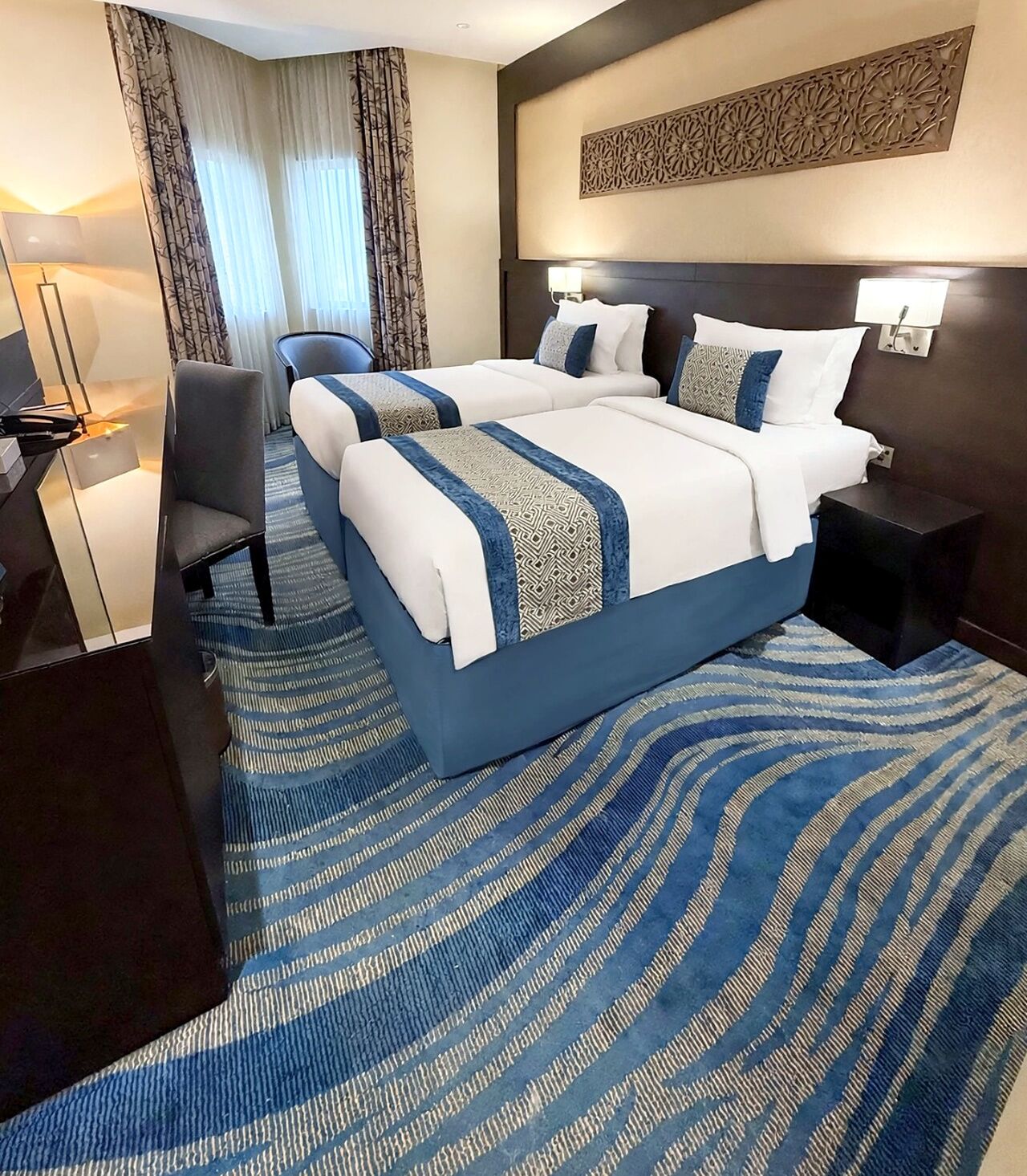 A hotel room with a large bed, a desk, a chair, and a decorative wall panel.