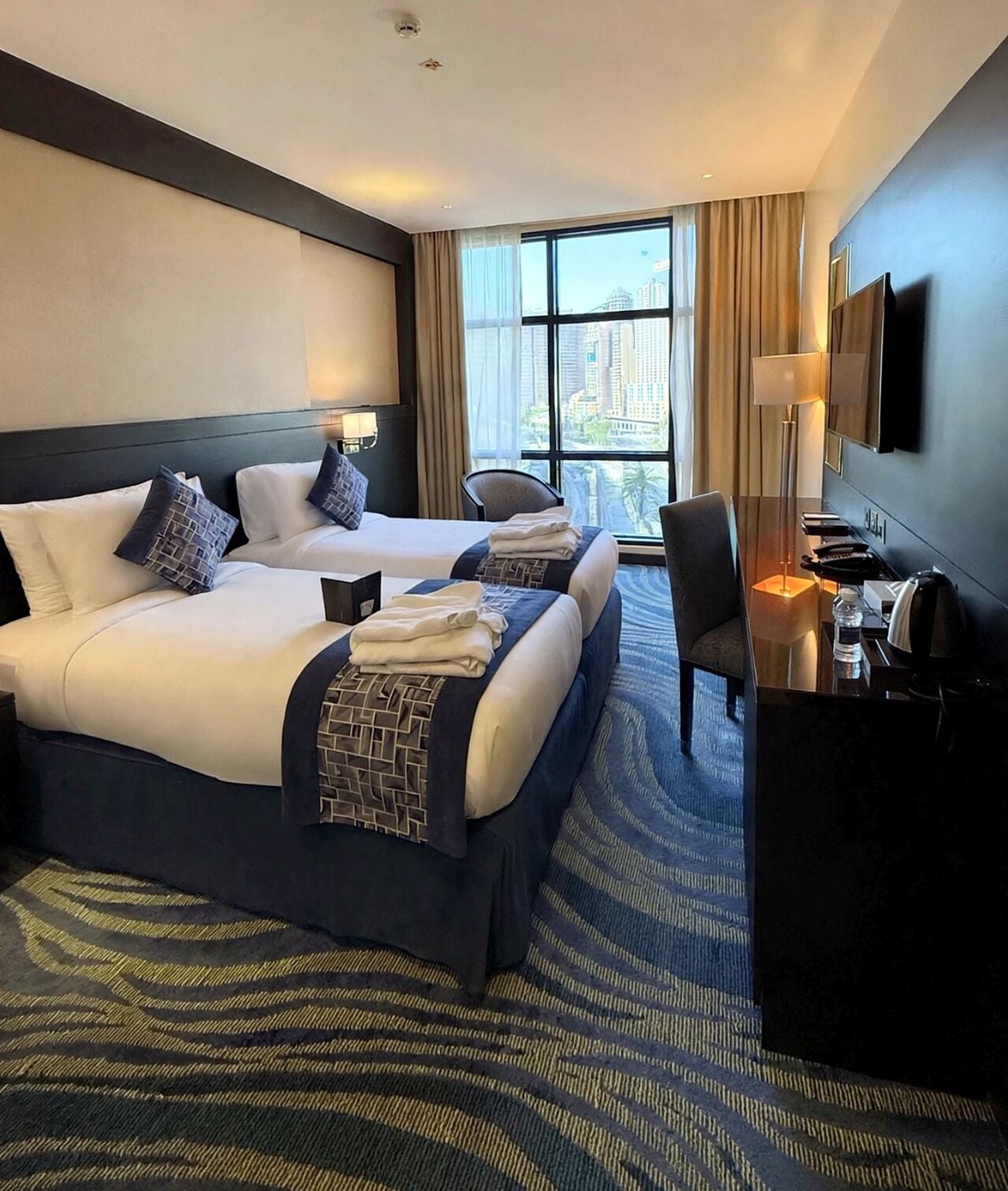 A hotel room with two beds, a desk, a chair, a flat-screen TV, and a large window offering a city view.