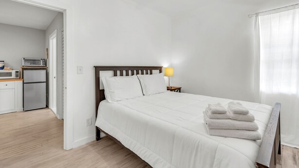 Een slaapkamer met een bed, kussens, een bedlampje, een stapel handdoeken en uitzicht op een keuken met apparatuur.