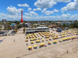 Ein Strand mit vielen Liegestühlen und Sonnenschirmen, ein Parkplatz und ein Gebäude mit flachem Dach.