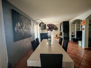 Zona de menjador amb taula llarga, cadires, un ram de flors, un quadre a la paret i un televisor al fons.