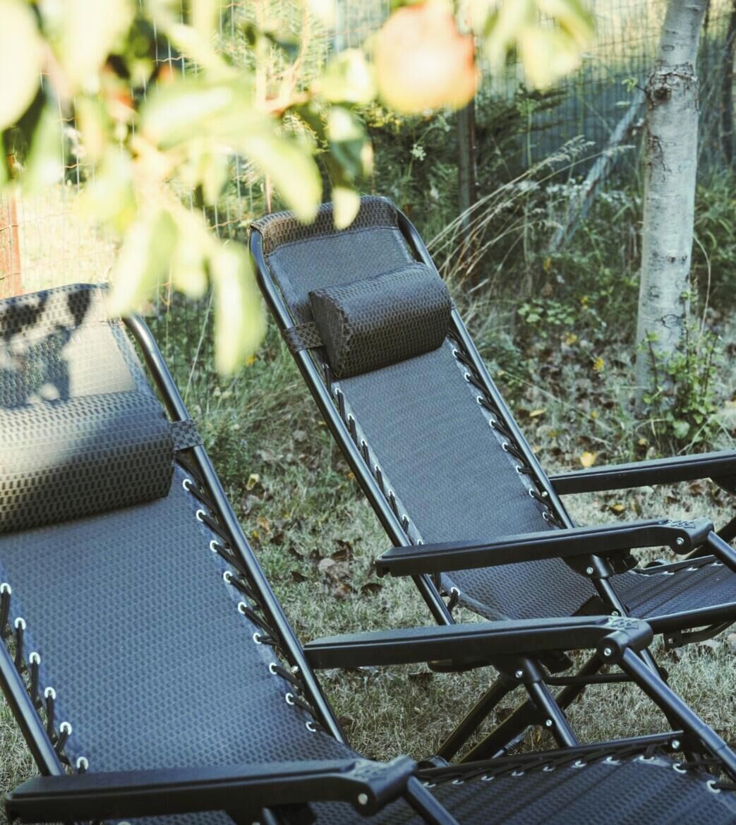 A row of lounge chairs with a mesh surface and a padded headrest, arranged outdoors with a fence and vegetation in the background.