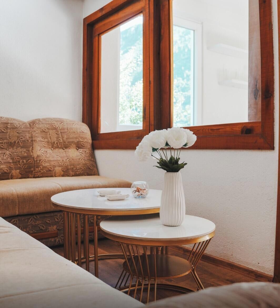 Een kamer met een bank, een ronde tafel met een bloevas, en een groot raam.