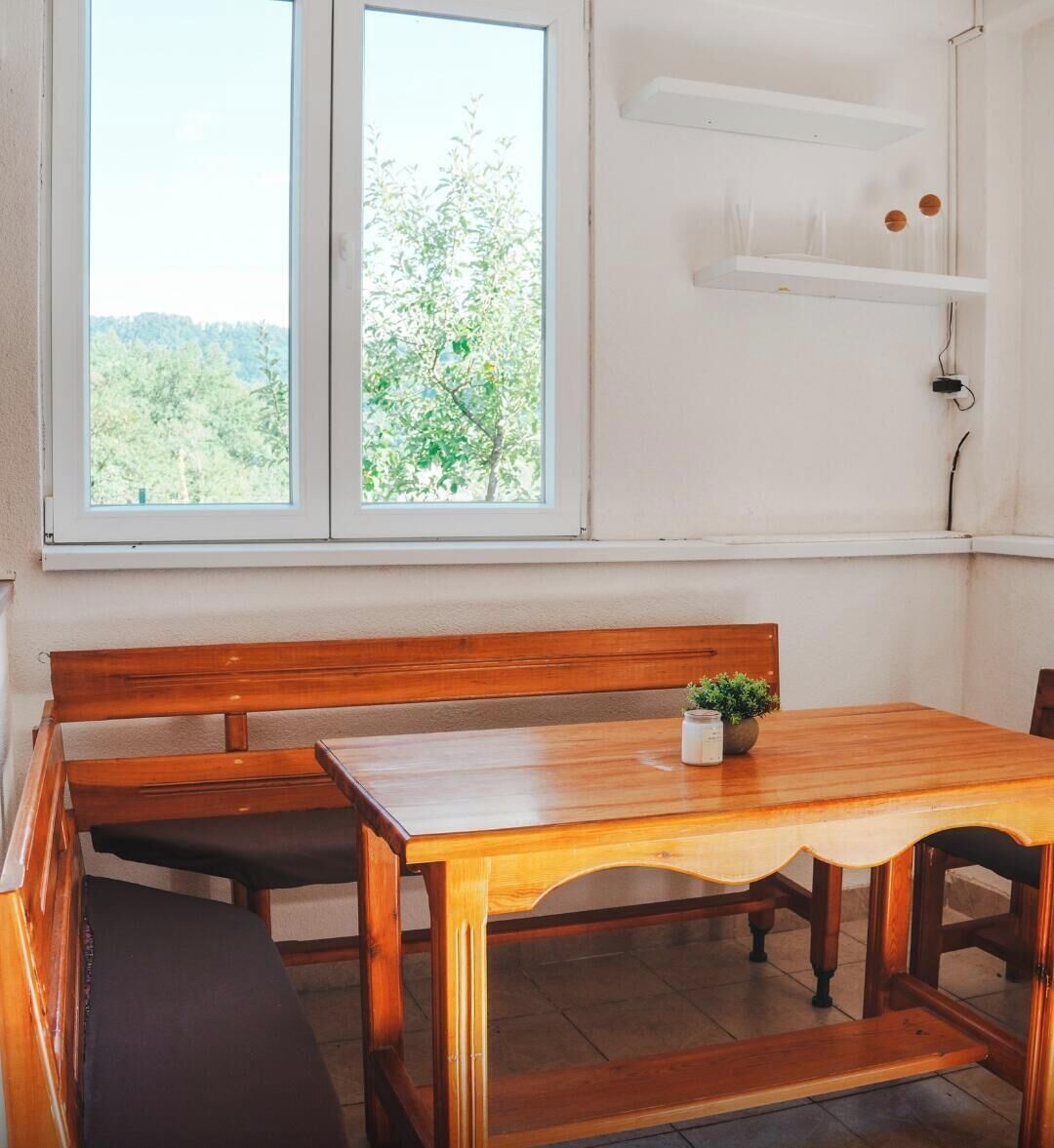 Une table à manger en bois, avec des bancs, une plante en pot et une vue sur la verdure par la fenêtre.