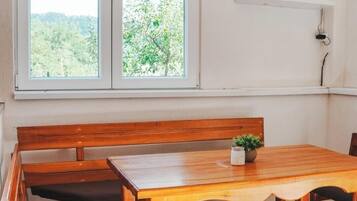 Une table à manger en bois, avec des bancs, une plante en pot et une vue sur la verdure par la fenêtre.