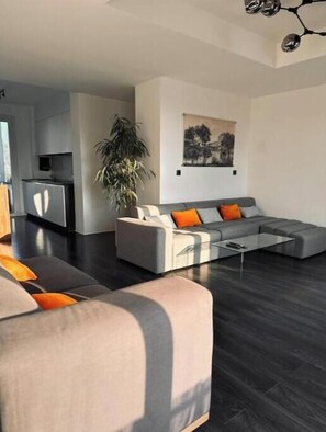Ein modernes Wohnzimmer mit einem Ecksofa, einem Glastisch und einem Essbereich, im Hintergrund ist eine Küche zu sehen.
