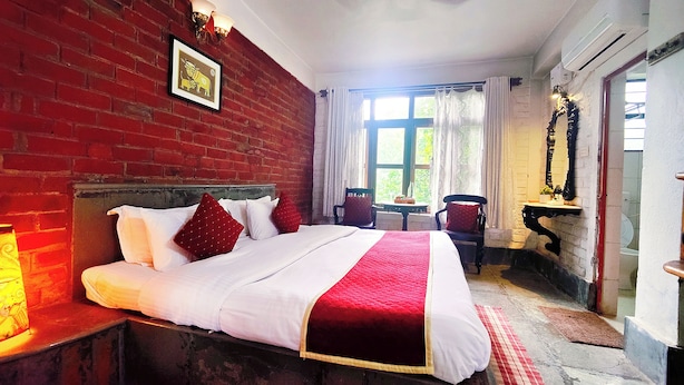 Une chambre avec un mur en briques, un lit avec une literie blanche et rouge, une chaise, une table et une fenêtre avec des rideaux.