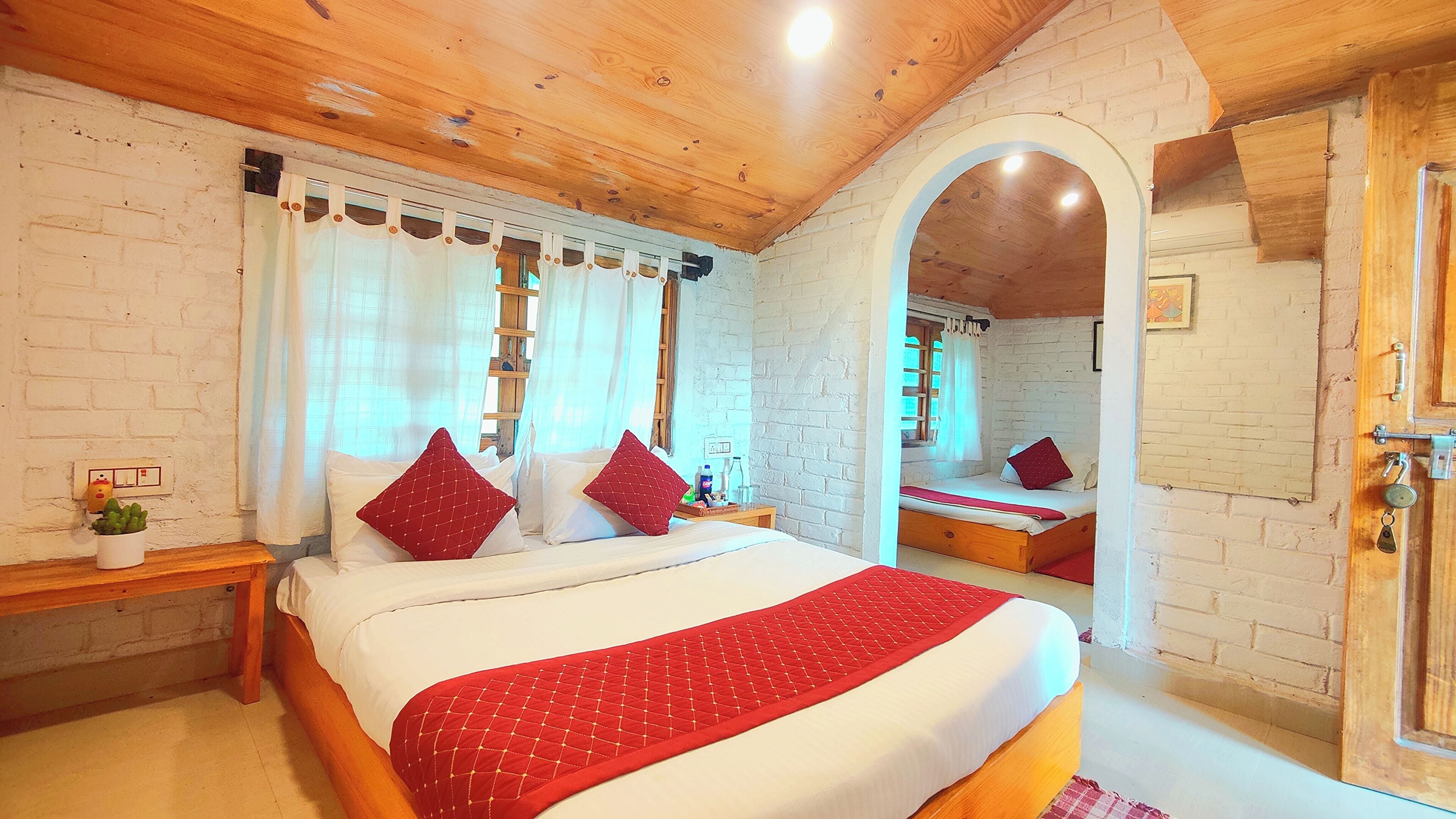 A bedroom with a wooden ceiling, white brick walls, a bed with red bedding, a bedside table with a plant, and a mirror on the wall.
