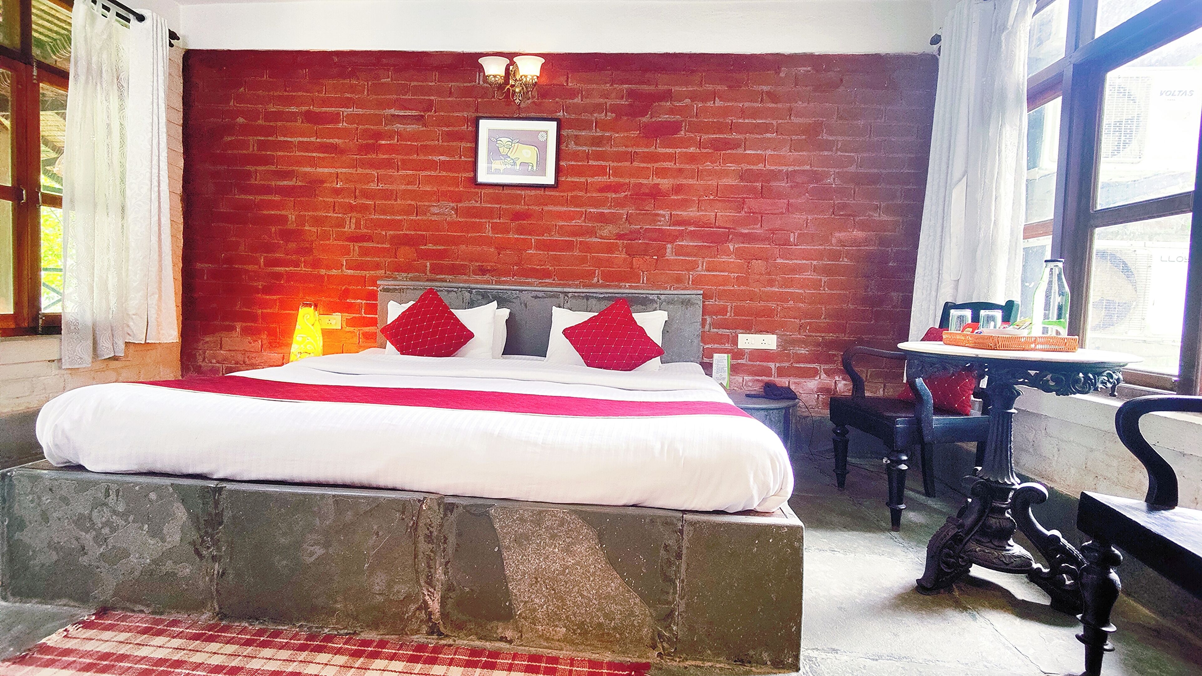 Una habitación con pared de ladrillo, una cama con ropa de cama blanca y almohadas rojas, una mesita con una botella y una silla.