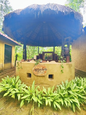 A tea stall with a thatched roof, surrounded by greenery.