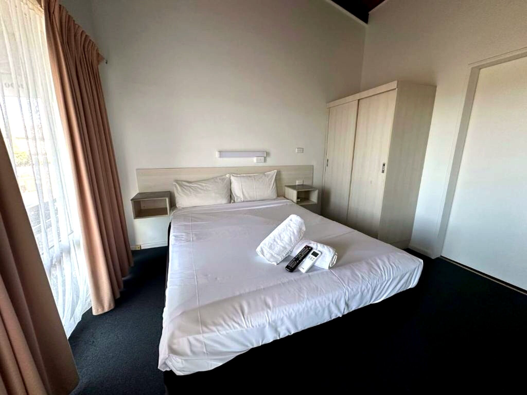 Habitación de hotel con una cama grande, una mesita de noche, un armario y una ventana con cortinas.