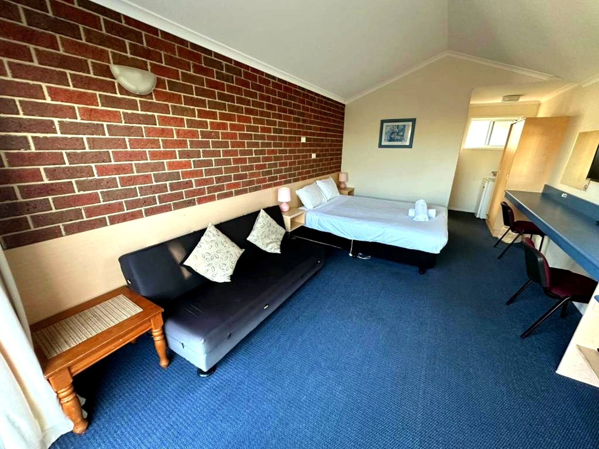 A room with a brick wall, a bed, a sofa, a small table, and a desk with chairs.