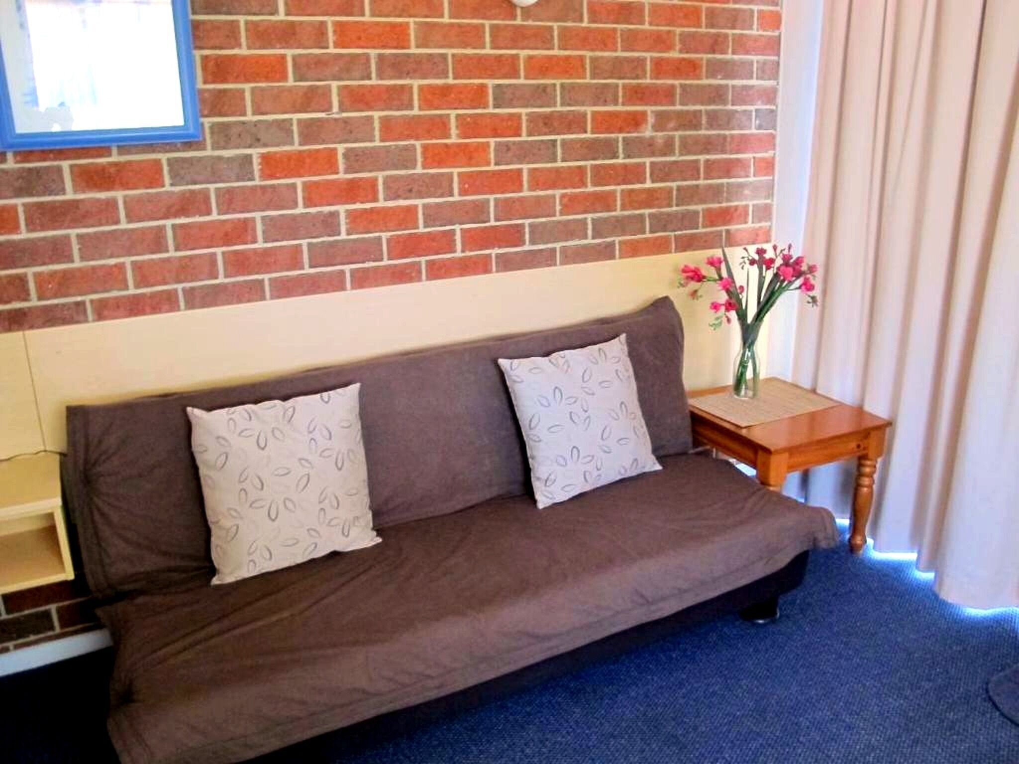A brown sofa with patterned pillows, a small wooden side table with a vase of flowers, and a brick wall.