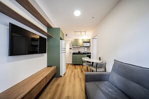 A modern living room with a flat-screen TV, a grey sofa, a wooden bench, and a kitchen area with green cabinets and a microwave. - LA CASA PÁSSAROS (São Paulo)