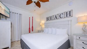 A hotel room with a large bed, a ceiling fan, a TV, a nightstand, and a wall sign that reads 'BEACH'. - Phoenix Viii 81105 1 Bedroom Condo (Orange Beach)