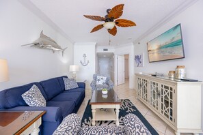 Ein Wohnzimmer mit blauem Sofa, gemusterten Kissen, einem Glastisch, Deckenventilator und einer Wanddekoration in Form eines Hais.