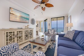 Ein Wohnzimmer mit Deckenventilator, einer Couch, einem Couchtisch und Blick auf den Ozean durch große Fenster.