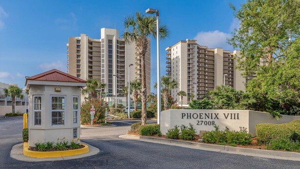 Uma entrada com portão e placa indicando 'PHOENIX VIII 27008', edifícios residenciais altos e uma pequena cabine de segurança.