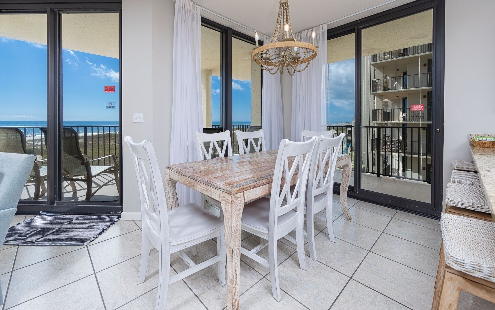 A dining area with a wooden table and white chairs, a chandelier, and a view of the sea through large glass doors.