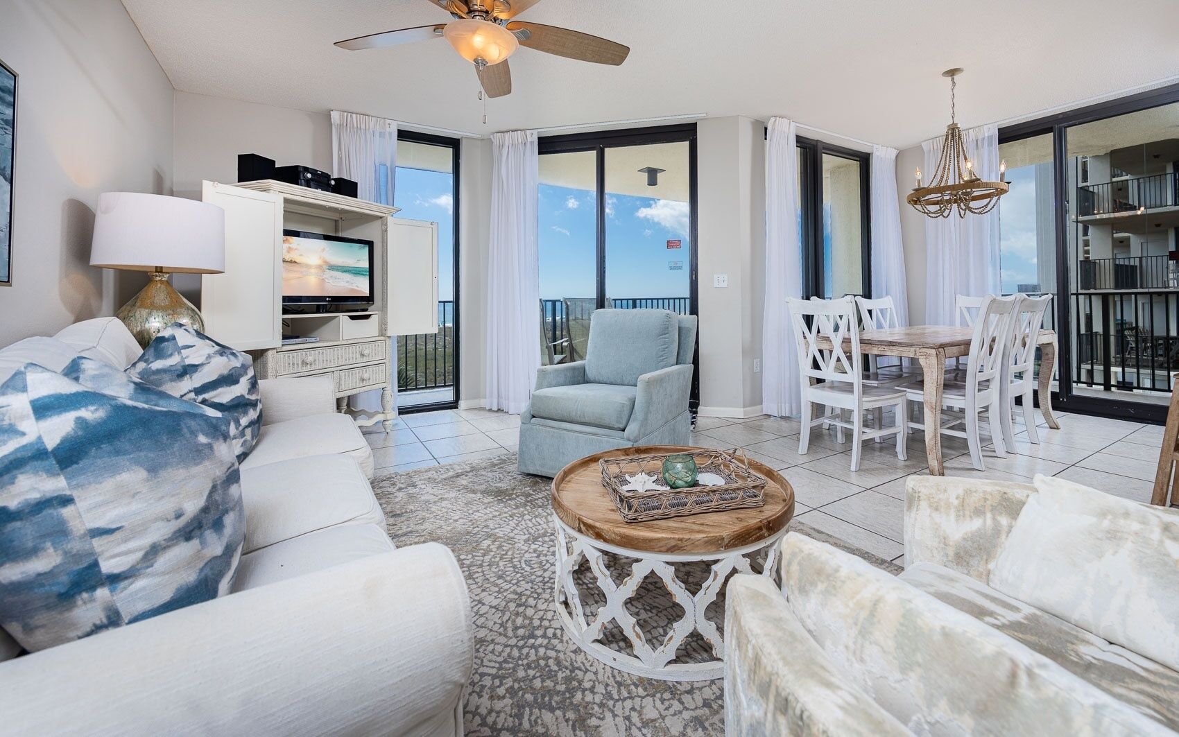 A spacious living area with a ceiling fan, a television, a dining table with chairs, and a balcony with a view.