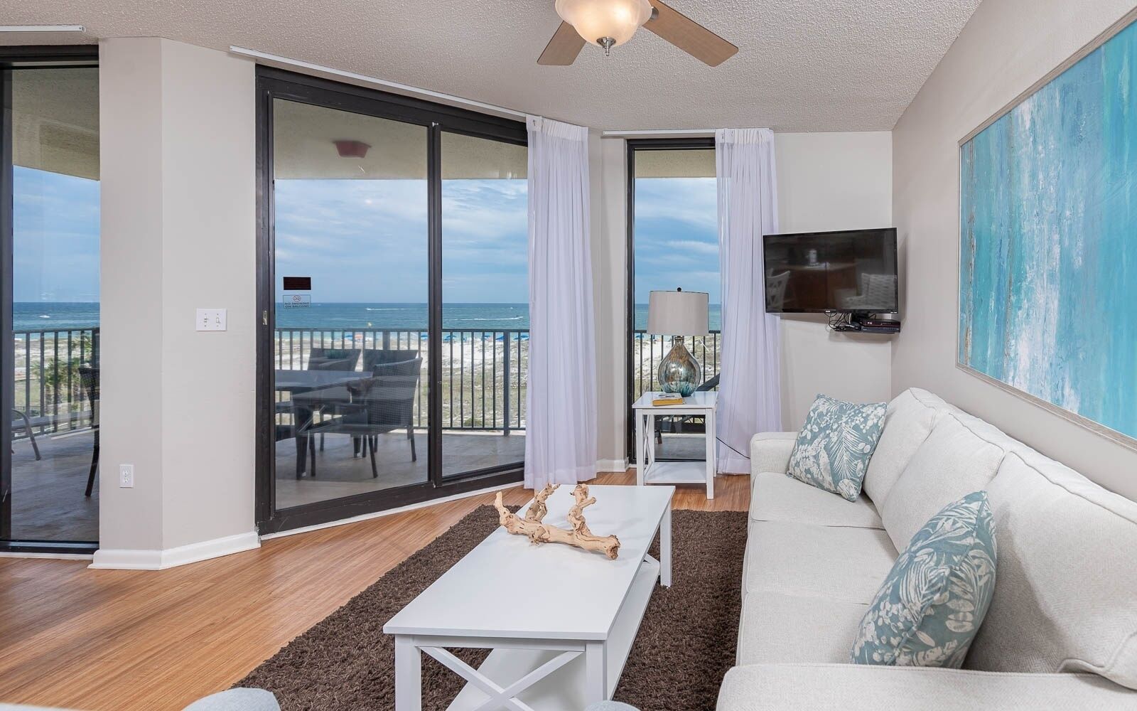A living room with a view of the beach, a ceiling fan, a flat-screen TV, and a white couch.