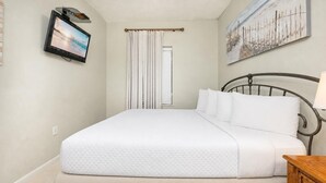 Ein ordentlich bezogenes Bett mit einer gesteppten Decke, ein an der Wand befestigter Fernseher, ein Gemälde über dem Bett und ein Fenster mit Vorhängen.