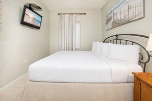 A neatly made bed with a quilted cover, a television mounted on the wall, a painting above the bed, and a window with curtains. - Phoenix Vii 71508 3 Bedroom Condo (Orange Beach)