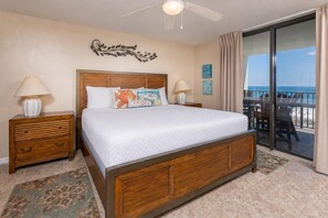 A hotel room with a large bed, wooden headboard, two bedside tables with lamps, a decorative wall piece, and a view of the beach through a sliding glass door. - Phoenix Vii 7708 3 Bedroom Condo (Orange Beach)