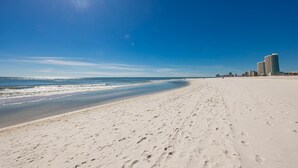 Ein weiter Sandstrand mit Fußabdrücken, strahlend blauer Himmel und entfernte Gebäude.