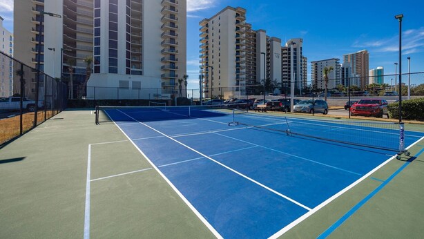 A tennis court with a net, surrounded by high-rise buildings and parked cars. - Phoenix Viii 8706 3 Bedroom Condo (Orange Beach)