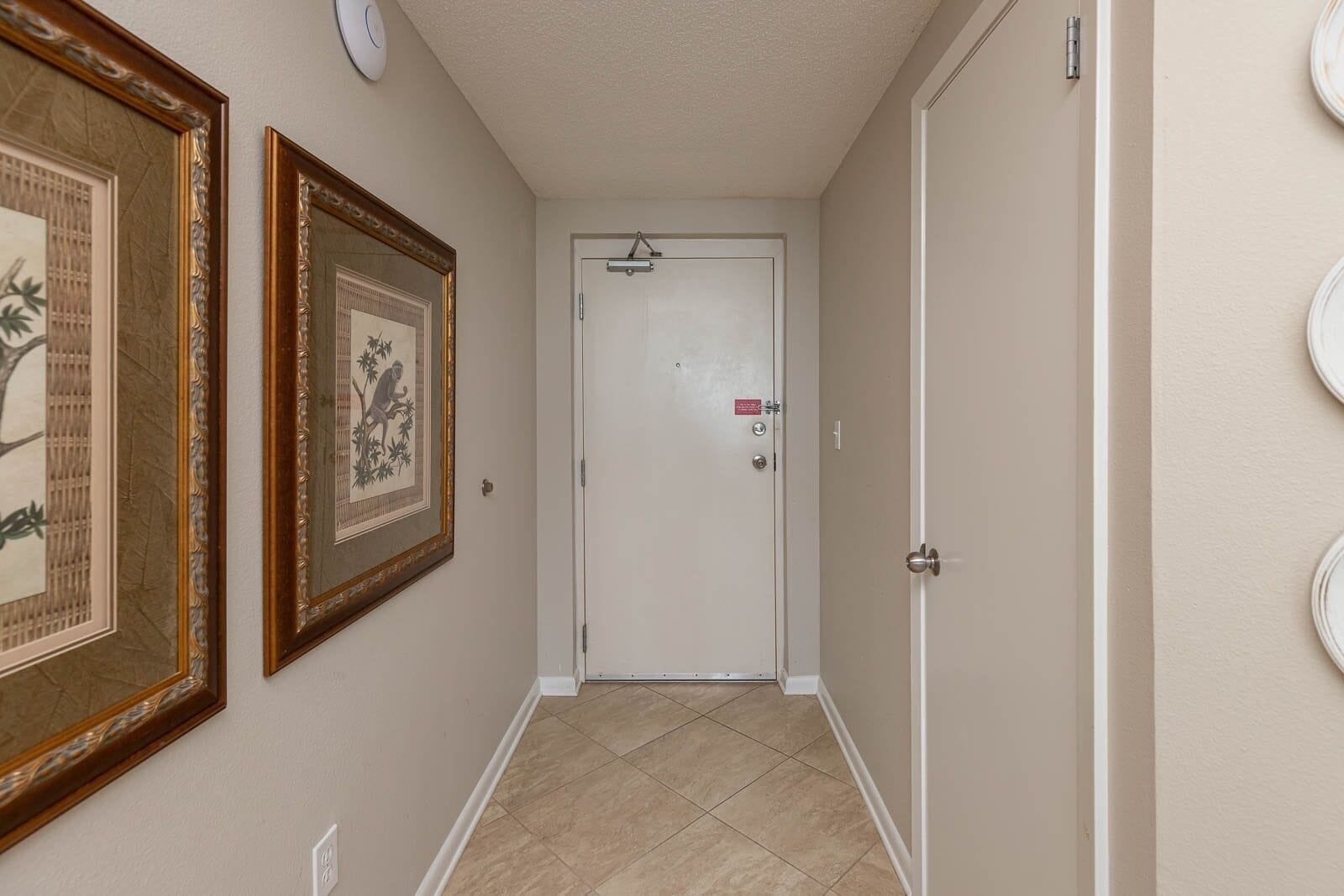 A hallway with framed artwork on the walls, a white door with a red sign, and a tiled floor.