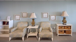 A waiting area with two patterned armchairs, a small table, and a side table with a lamp. There are framed pictures on the wall and a bookshelf with books. - Phoenix Viii 81003 3 Bedroom Condo (Orange Beach)
