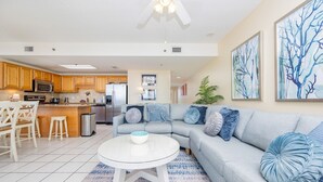 A modern kitchen with wooden cabinets, stainless steel appliances, and a dining area with white chairs. - Phoenix Viii 8701 3 Bedroom Condo (Orange Beach)
