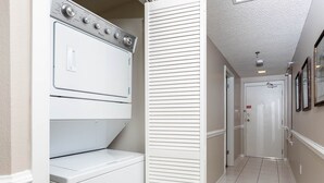 A laundry room with stacked washers and dryers, a door leading to a hallway, and a tiled floor. - Phoenix Viii 81002 2 Bedroom Condo (Orange Beach)