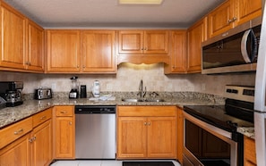 A kitchen with wooden cabinets, granite countertops, a dishwasher, a coffee maker, a toaster, a blender, and a microwave. - Phoenix Viii 81002 2 Bedroom Condo (Orange Beach)